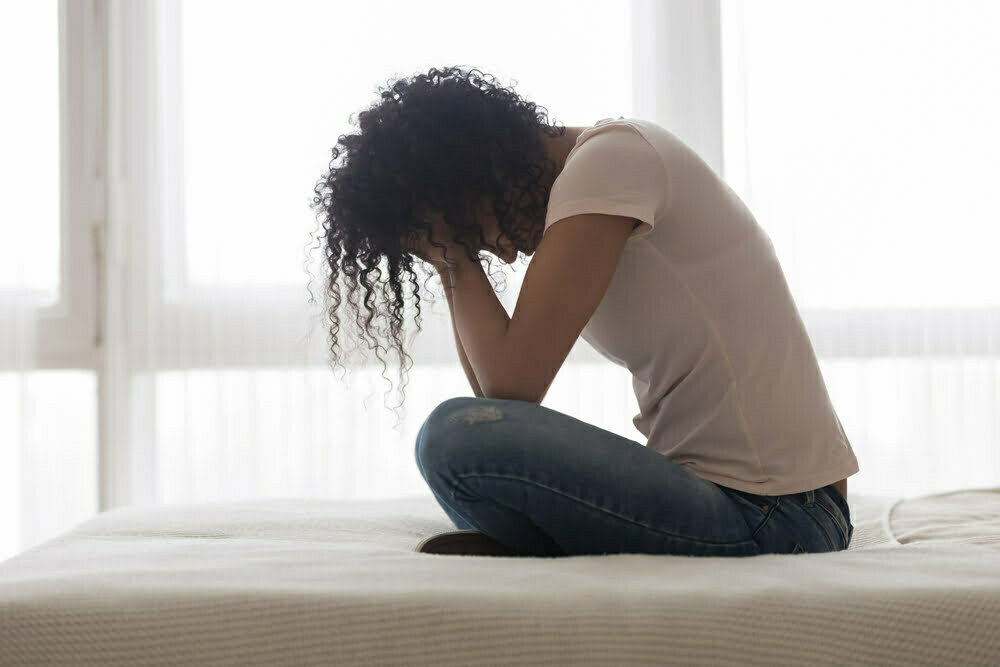 African American woman sitting on her bed, holding her head, and struggling with anxiety